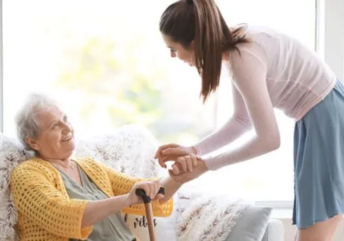 A young female caregiver gently assisting an elderly woman with a walking stick, both smiling warmly in a bright, cozy home setting — representing compassionate support by a local NDIS provider.