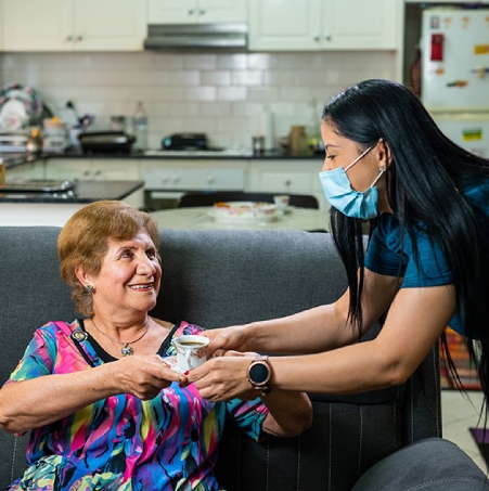 Carer in a mask serving coffee to smiling older woman for In-home support services